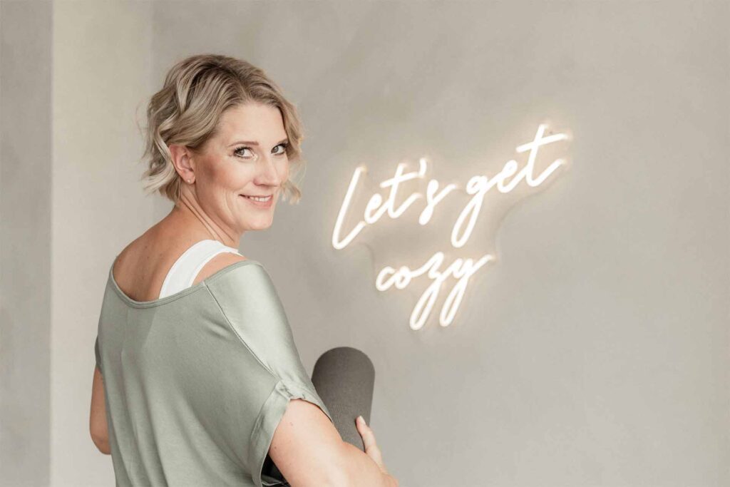 Meditation: Julia Jäger blickt lächelnd über die Schulter und hat eine Yogamatte unter dem Arm