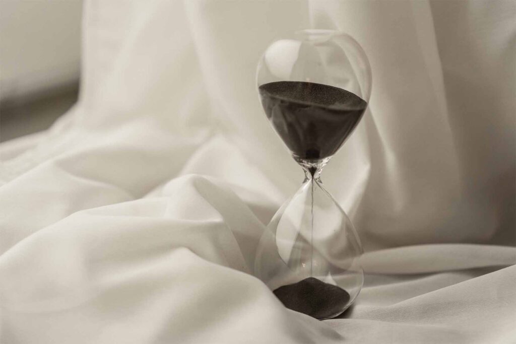 Was ist Achtsamkeit/ Mindfulness?: Sanduhr mit schwarzem Sand steht in weißen Tüchern